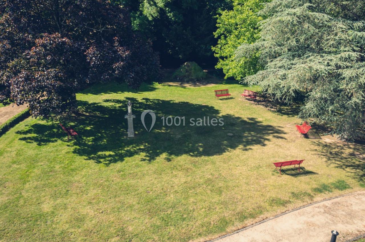 Vue d'un jardin avec pelouse, arbres, bancs rouges et une statue au centre, sous un ciel dégagé.