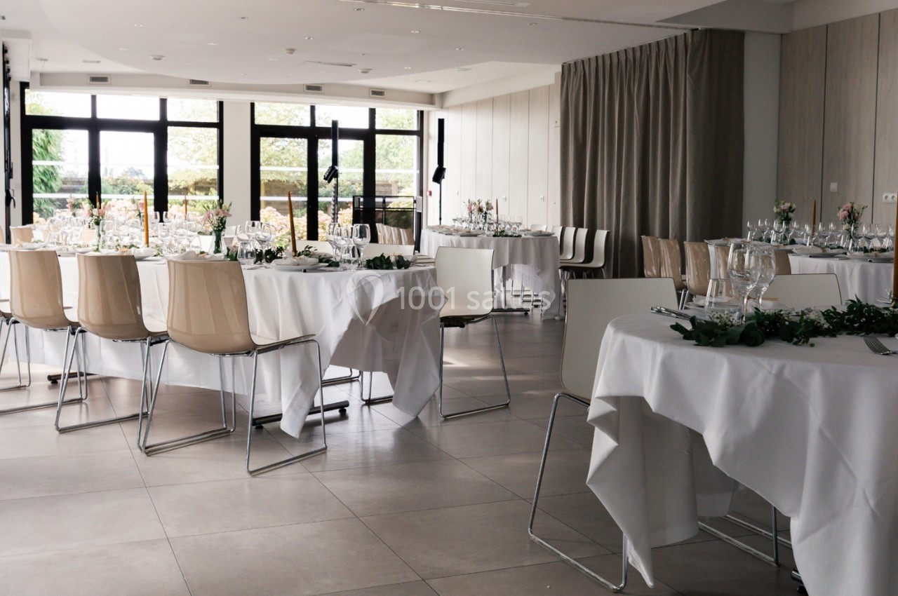 Salle de réception lumineuse avec tables dressées, nappes blanches et décoration florale, vue sur l'extérieur.