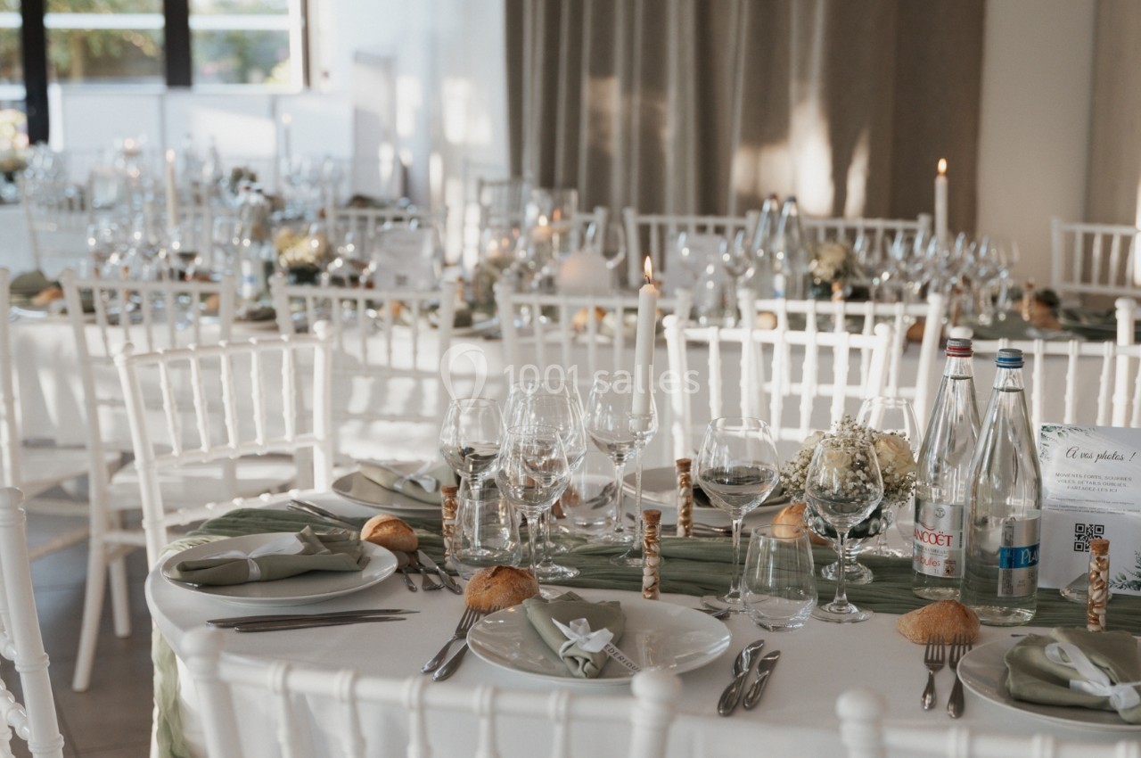Salle de réception décorée avec des tables dressées, nappes blanches, verres, bougies et chaises blanches.