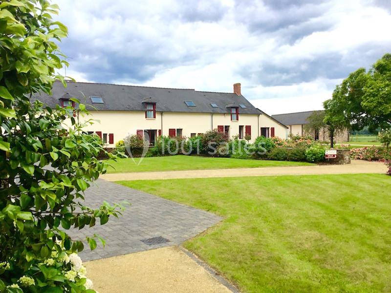 Bâtisse rurale avec façade blanche, volets rouges, jardin entretenu et allée pavée sous un ciel nuageux.