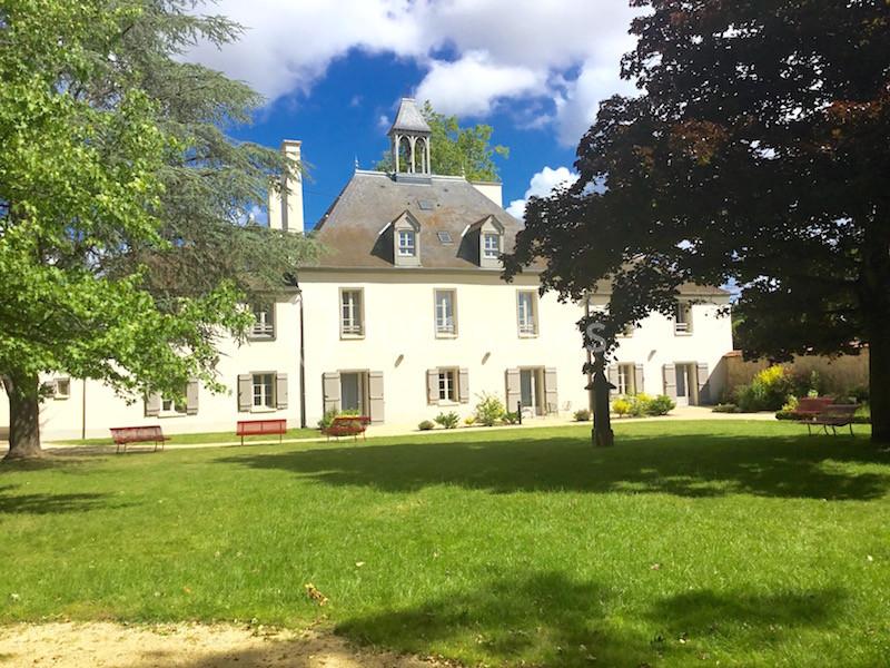 Grande maison blanche à deux étages avec un petit clocher, entourée d'arbres et d'une pelouse verdoyante.