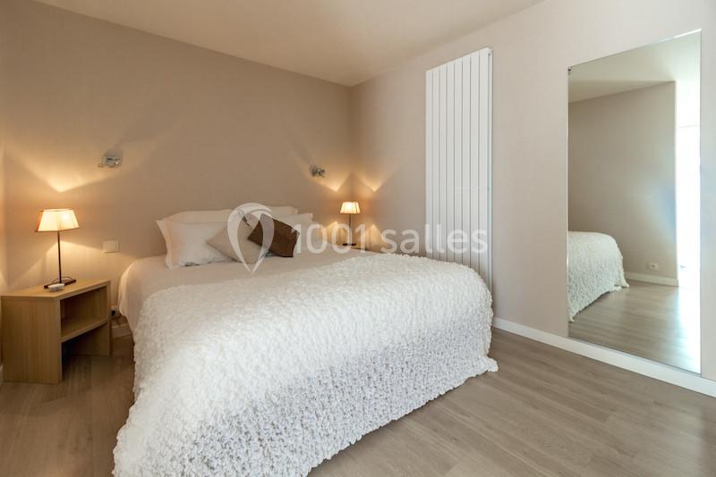 Chambre lumineuse avec lit double, couvre-lit texturé blanc, lampes de chevet et grand miroir sur un mur beige.