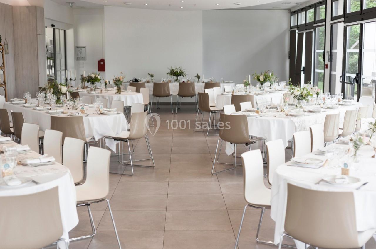 Salle lumineuse aménagée pour un événement, avec des tables rondes dressées et décorées de fleurs.