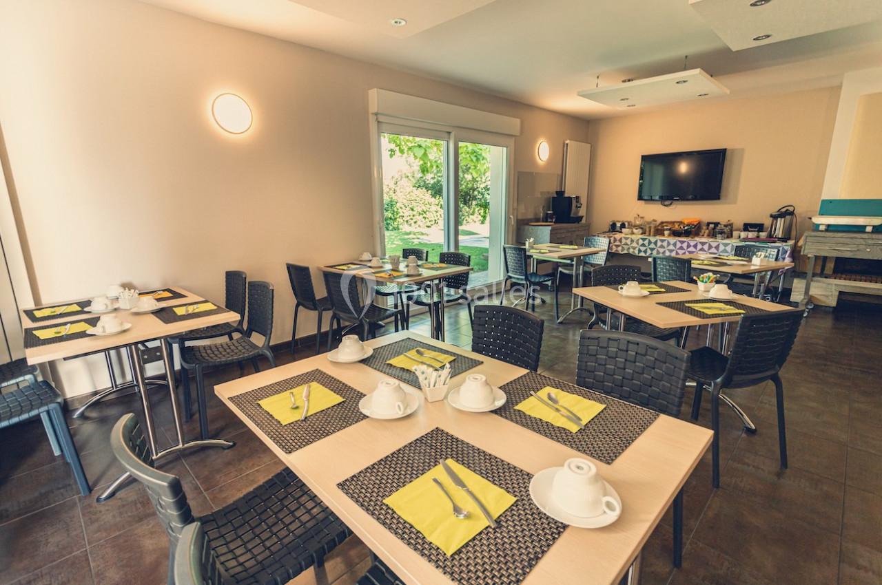 Salle lumineuse avec tables dressées pour le petit-déjeuner, chaises en osier, et vue sur un jardin à travers une baie…