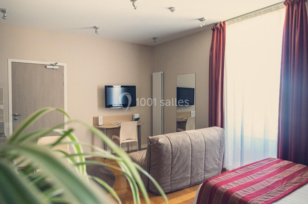 Chambre lumineuse avec lit, canapé, télévision murale, bureau et grande fenêtre avec rideaux rouges.