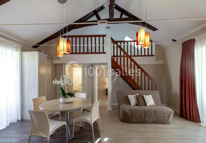 Salle à manger lumineuse avec table ronde, chaises blanches, canapé beige, poutres apparentes et escalier en bois.