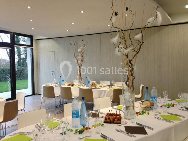 Salle décorée pour un repas, avec des tables dressées, centres de table ornés de branches et lumière naturelle.