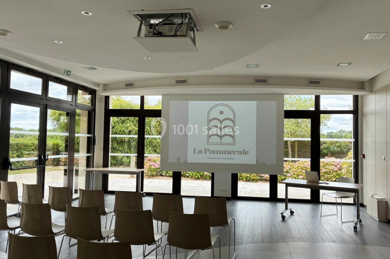 Salle de conférence lumineuse avec chaises alignées, écran de projection affichant un logo et vue sur un jardin extérieur.
