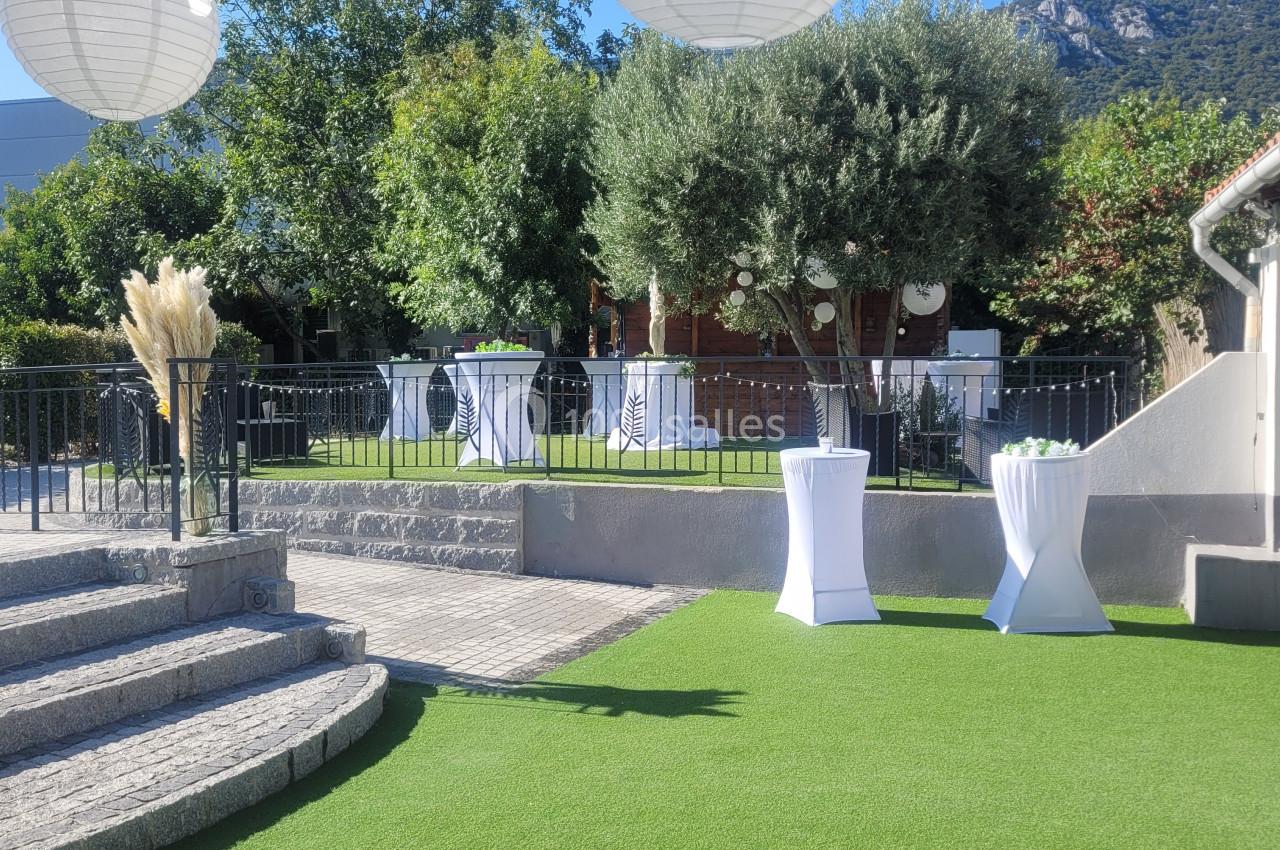 Jardin avec pelouse artificielle, lanternes suspendues, tables hautes blanches et vue sur des collines boisées.