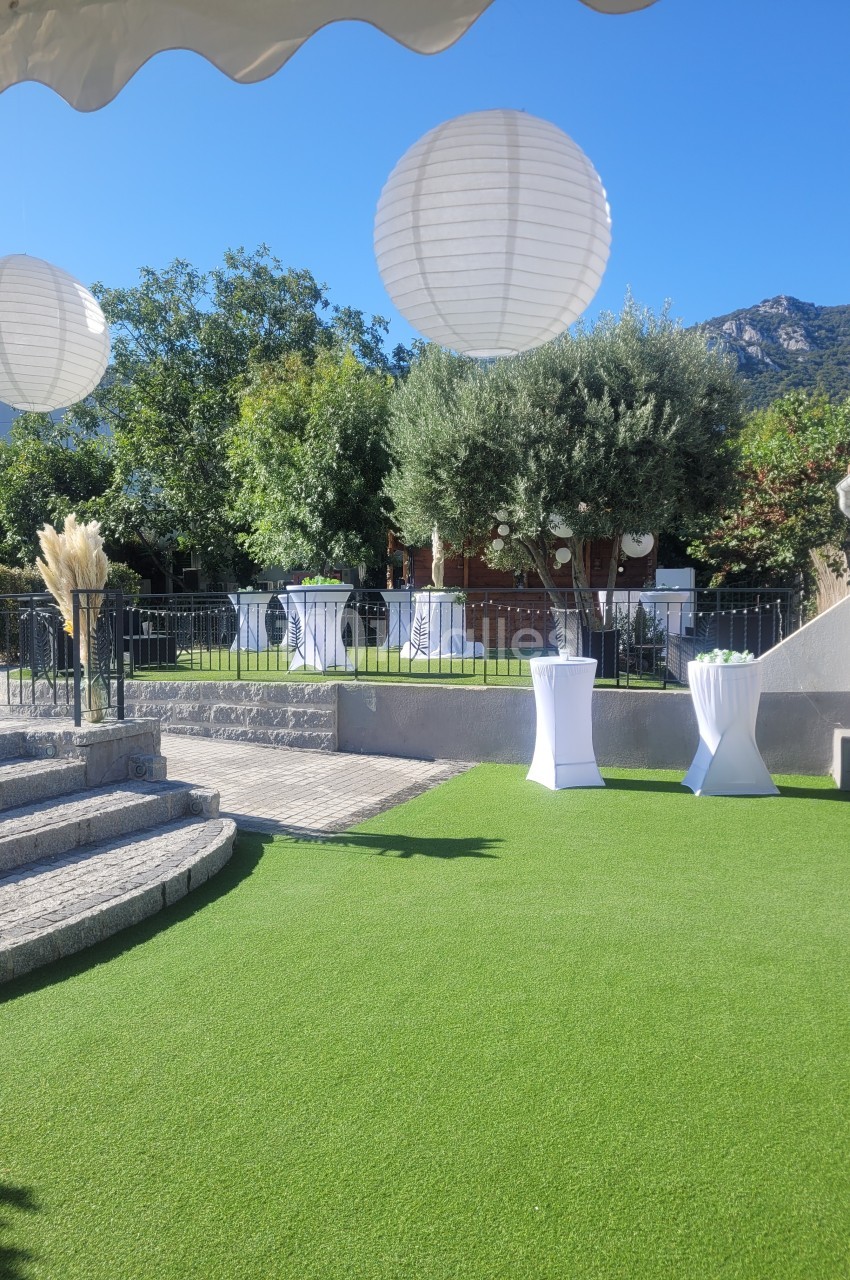 Jardin avec pelouse artificielle, lanternes suspendues, tables hautes blanches et vue sur des collines boisées.