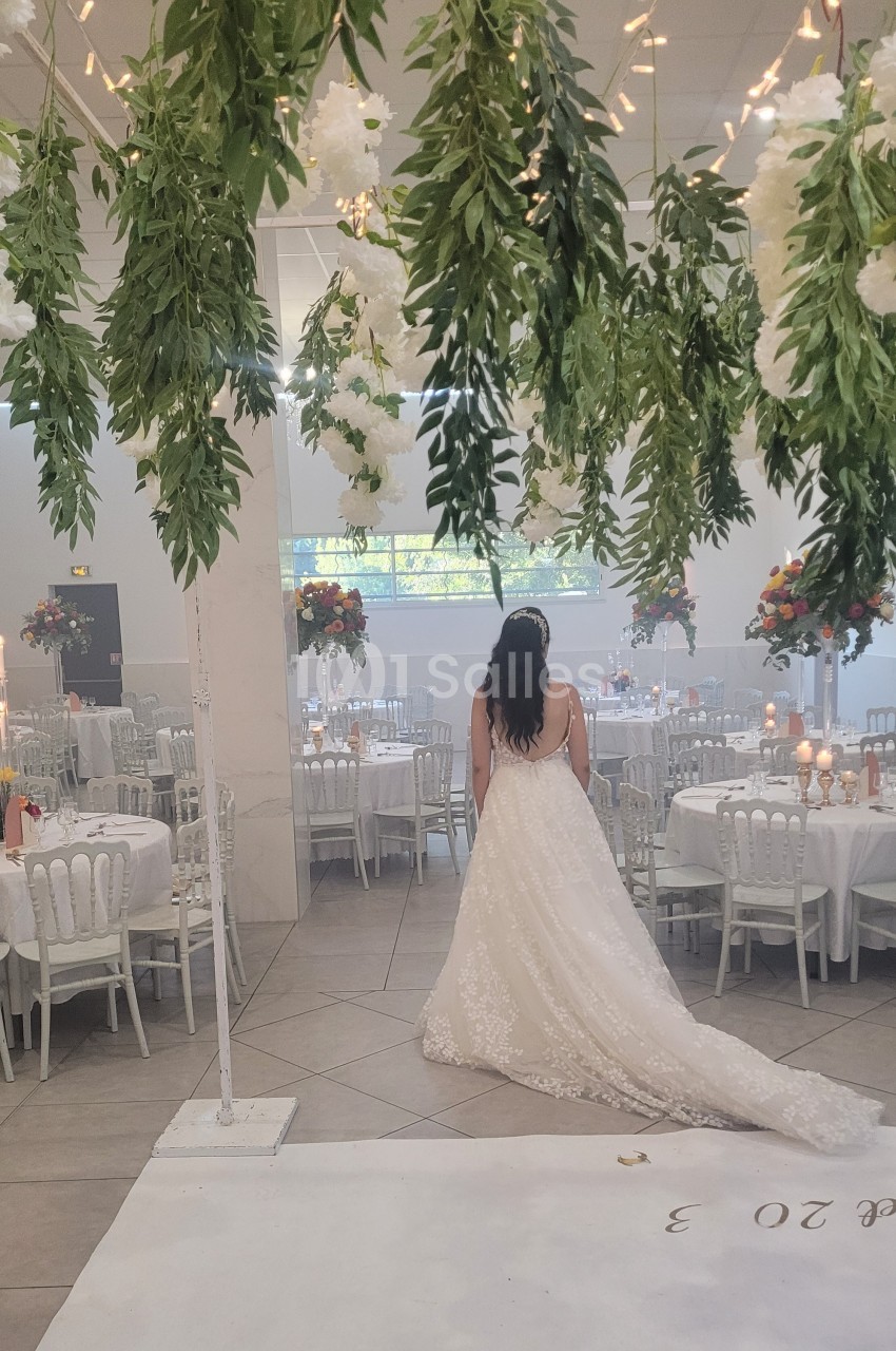 Une mariée en robe blanche regarde une salle de réception décorée de fleurs suspendues et de tables dressées.