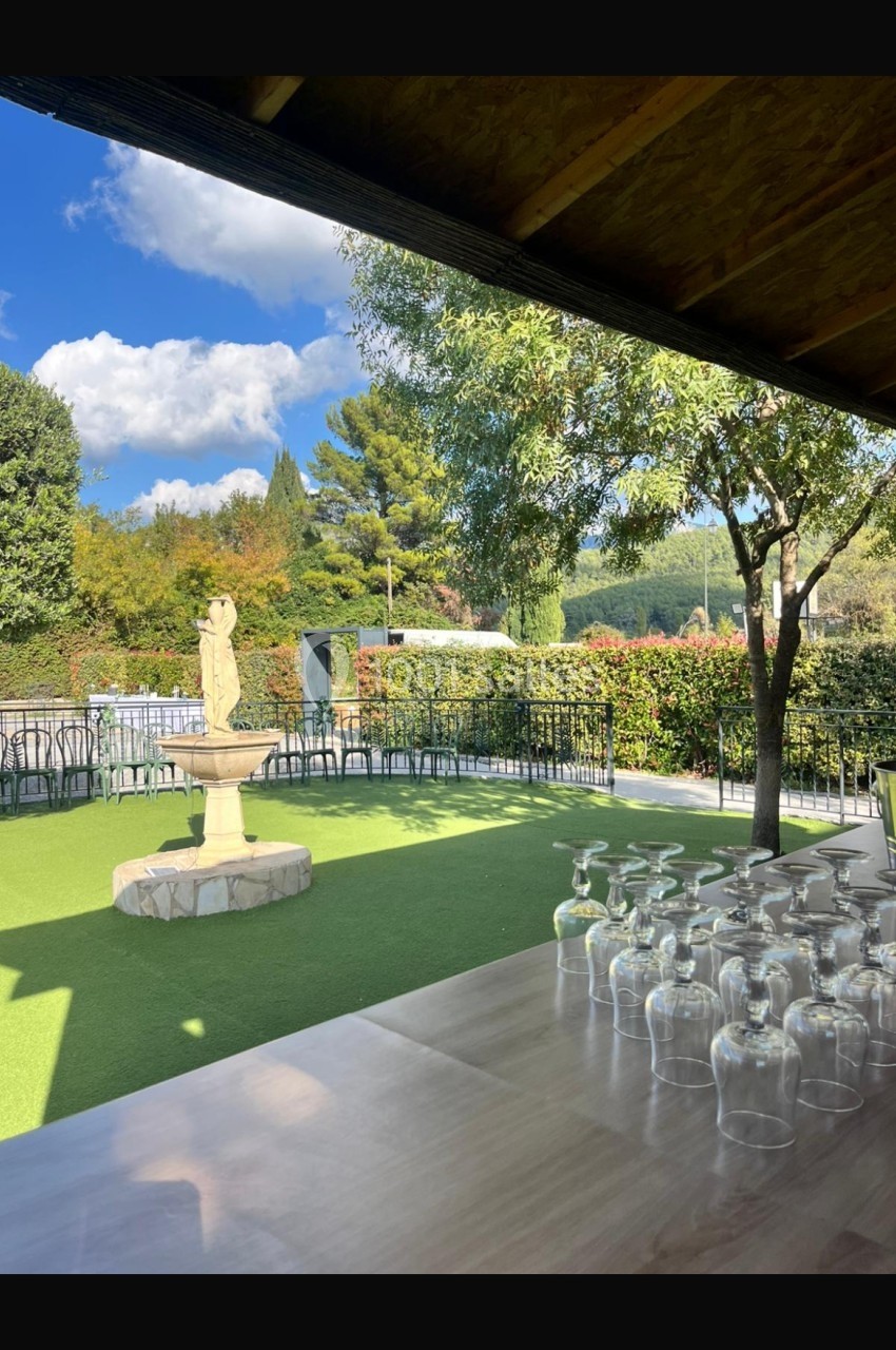 Fontaine en pierre dans un jardin verdoyant, vue depuis une table avec des verres retournés, sous un abri en bois.