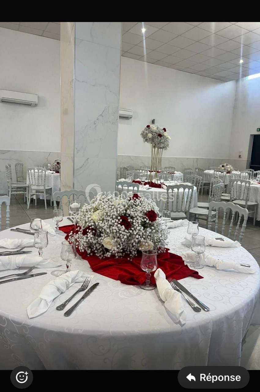 Salle de réception avec tables rondes décorées de nappes blanches, centres de table floraux rouges et blancs.