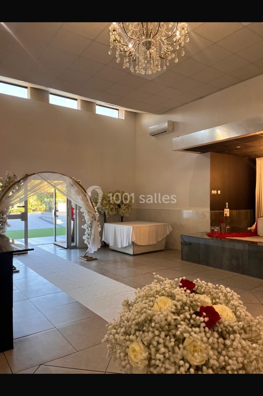 Salle de réception lumineuse avec arche décorative, fleurs blanches, lustre et table dressée près d'une baie vitrée.