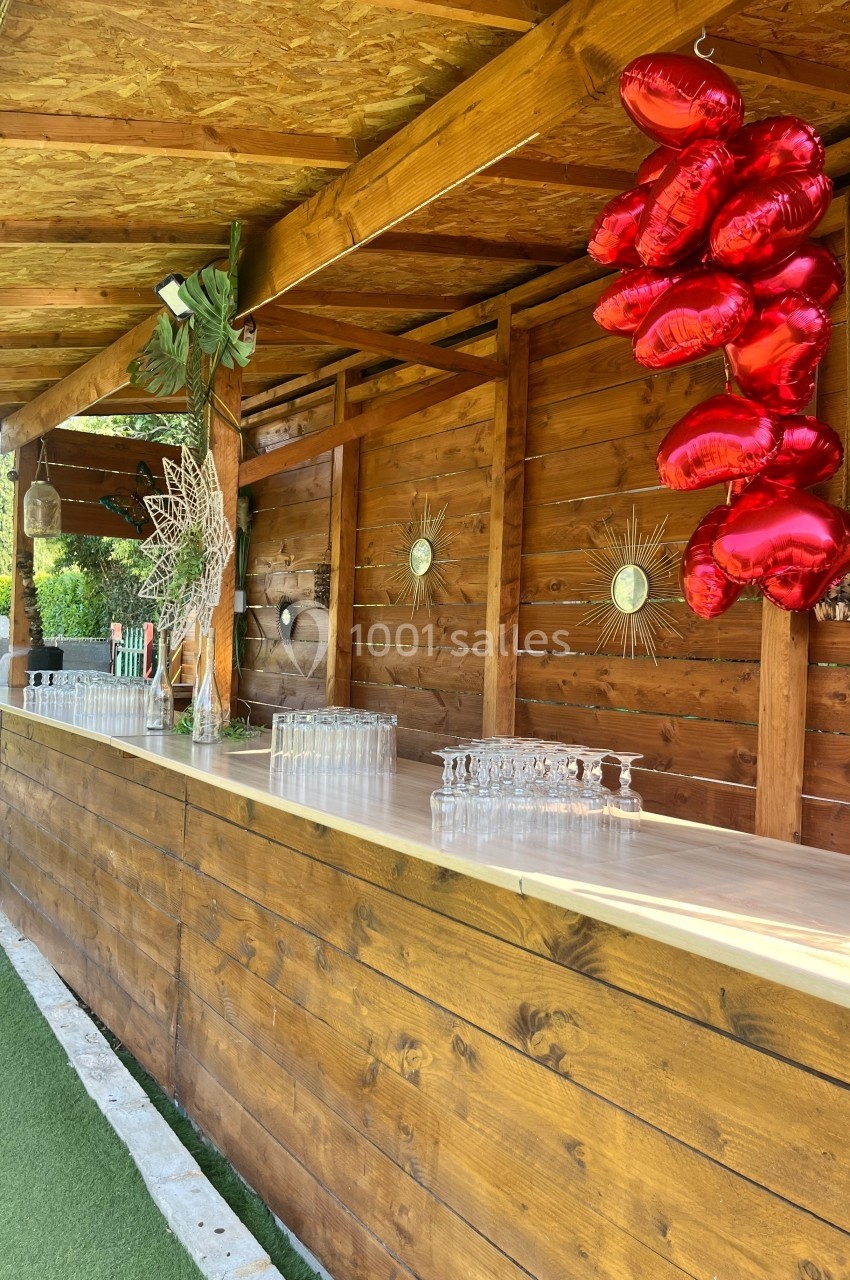 Comptoir en bois décoré de ballons rouges en forme de cœur et de verres alignés, situé en extérieur sous un auvent.