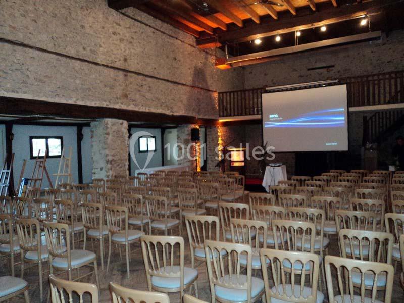 Salle de conférence avec des rangées de chaises en bois, un écran de projection et des murs en pierre apparente.