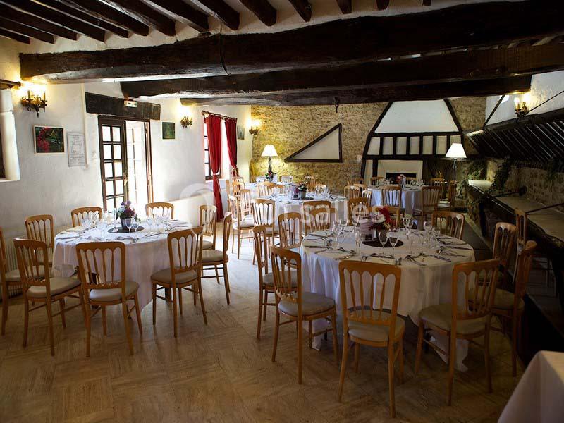 Salle de réception rustique avec tables rondes dressées, chaises en bois et poutres apparentes au plafond.