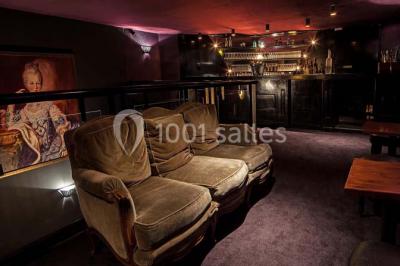 Personne en costume buvant un verre dans un bar faiblement éclairé, avec des lumières floues en arrière-plan.