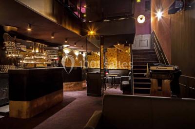 Personne en costume buvant un verre dans un bar faiblement éclairé, avec des lumières floues en arrière-plan.