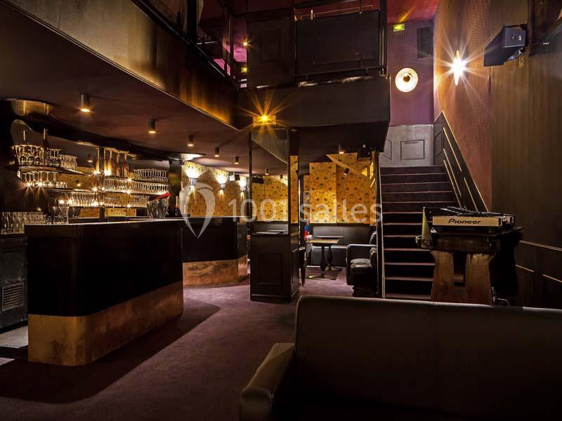 Intérieur d'un bar lounge avec éclairage tamisé, comptoir, tables, banquettes et escalier menant à un étage supérieur.
