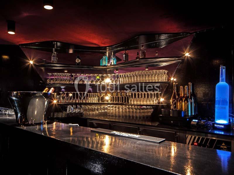 Bar moderne avec comptoir noir, étagères illuminées contenant des bouteilles et des verres, ambiance tamisée.