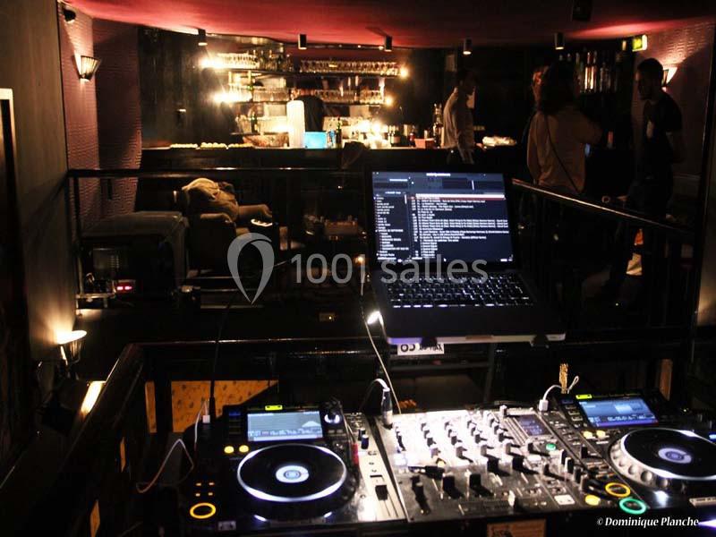 Poste de DJ avec platines et ordinateur dans un bar à l'éclairage tamisé, quelques personnes en arrière-plan.