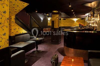 Personne en costume buvant un verre dans un bar faiblement éclairé, avec des lumières floues en arrière-plan.