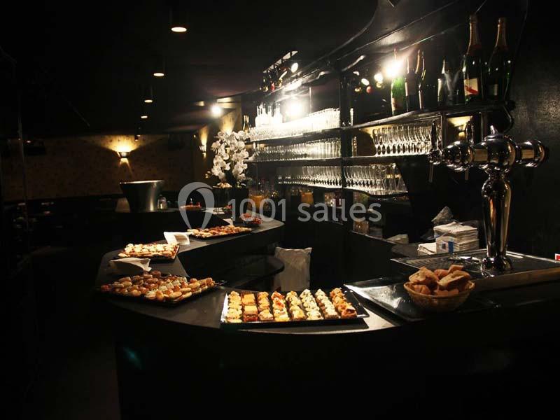 Bar sombre avec comptoir garni de plateaux de petits fours, verres alignés et bouteilles de champagne en arrière-plan.