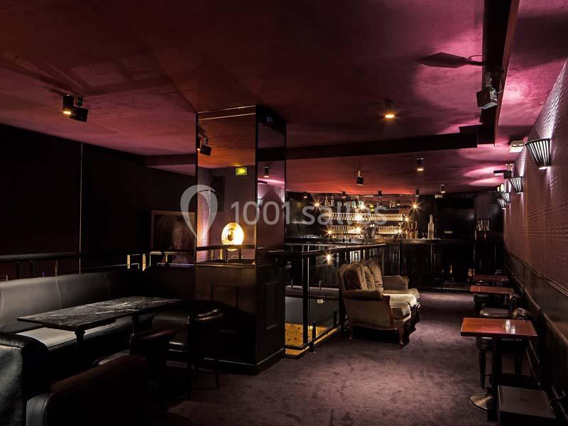 Salle de bar élégante avec éclairage tamisé, mobilier sombre et comptoir illuminé en arrière-plan.
