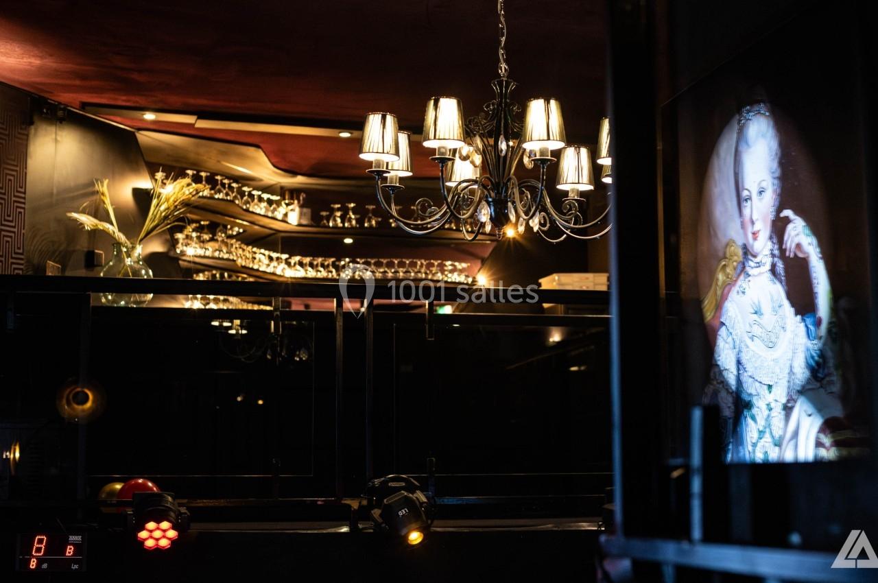 Un bar élégant avec des verres alignés, un lustre suspendu et un portrait éclairé en arrière-plan.