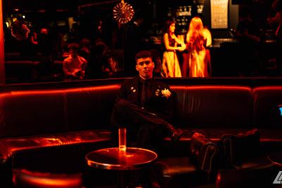 Personne en costume buvant un verre dans un bar faiblement éclairé, avec des lumières floues en arrière-plan.