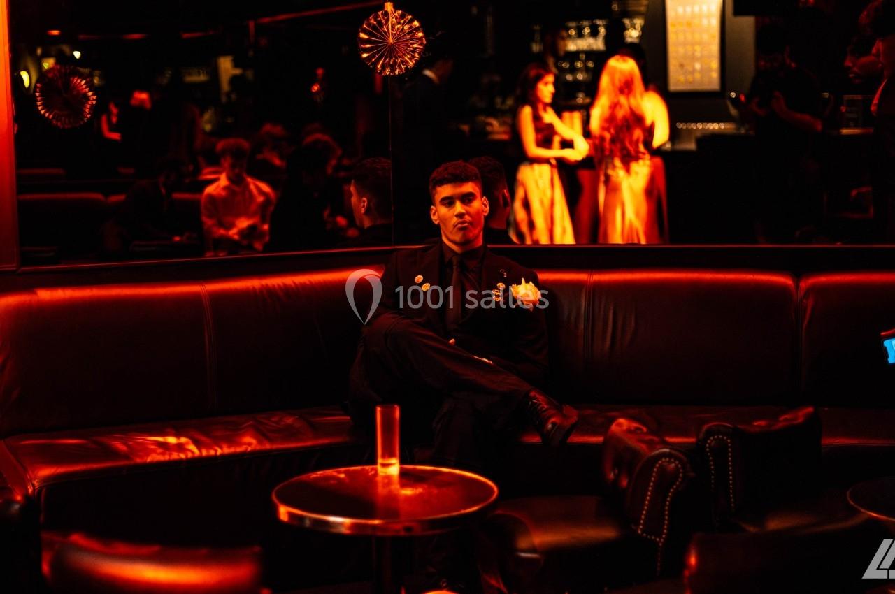 Un homme assis seul sur un canapé dans un espace sombre éclairé par des lumières rouges, avec des personnes en arrière-plan.