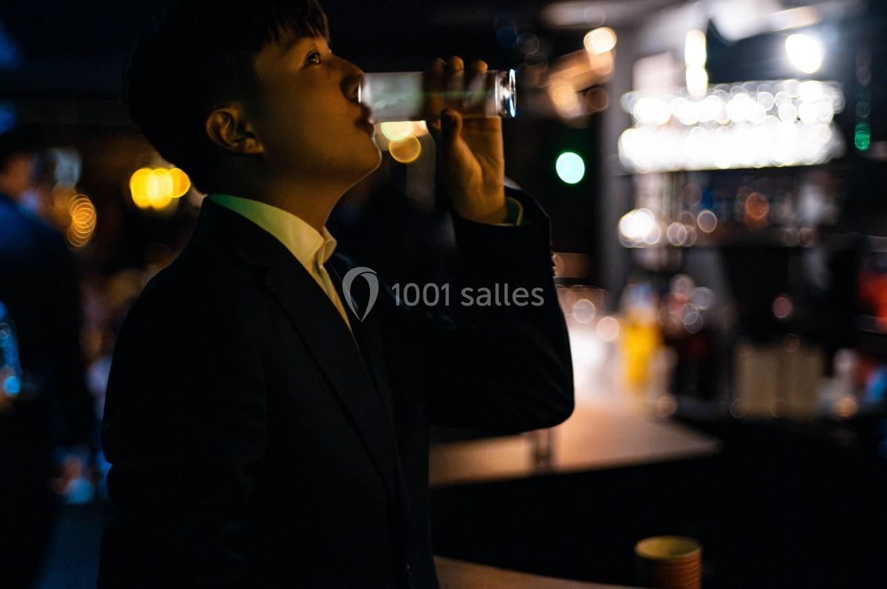 Personne en costume buvant un verre dans un bar faiblement éclairé, avec des lumières floues en arrière-plan.