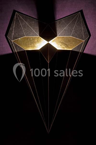 Structure géométrique en forme de masque avec des reflets dorés, éclairée sur un fond sombre.