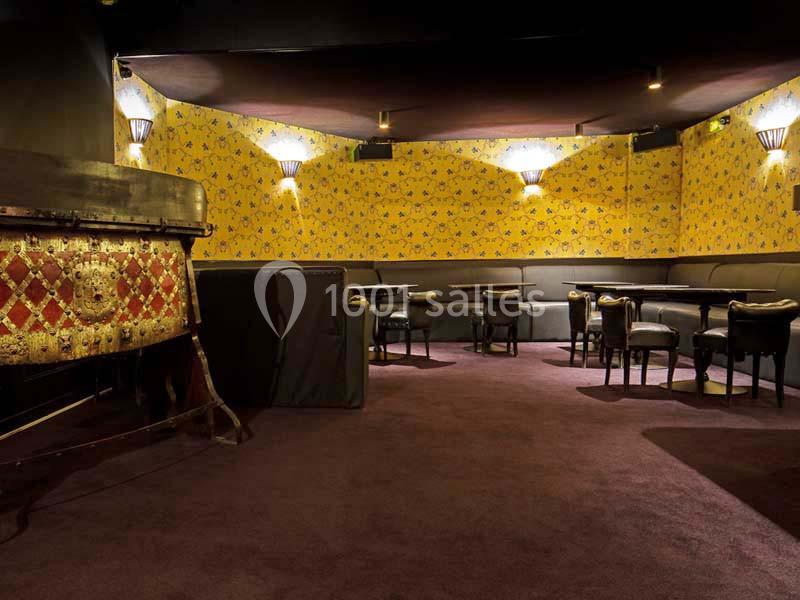 Salle avec moquette violette, murs jaunes ornés de motifs, tables rondes et chaises noires dans une ambiance feutrée.