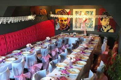 Salle décorée pour un événement avec longues tables dressées, nappes blanches, nœuds roses et portraits accrochés au mur.