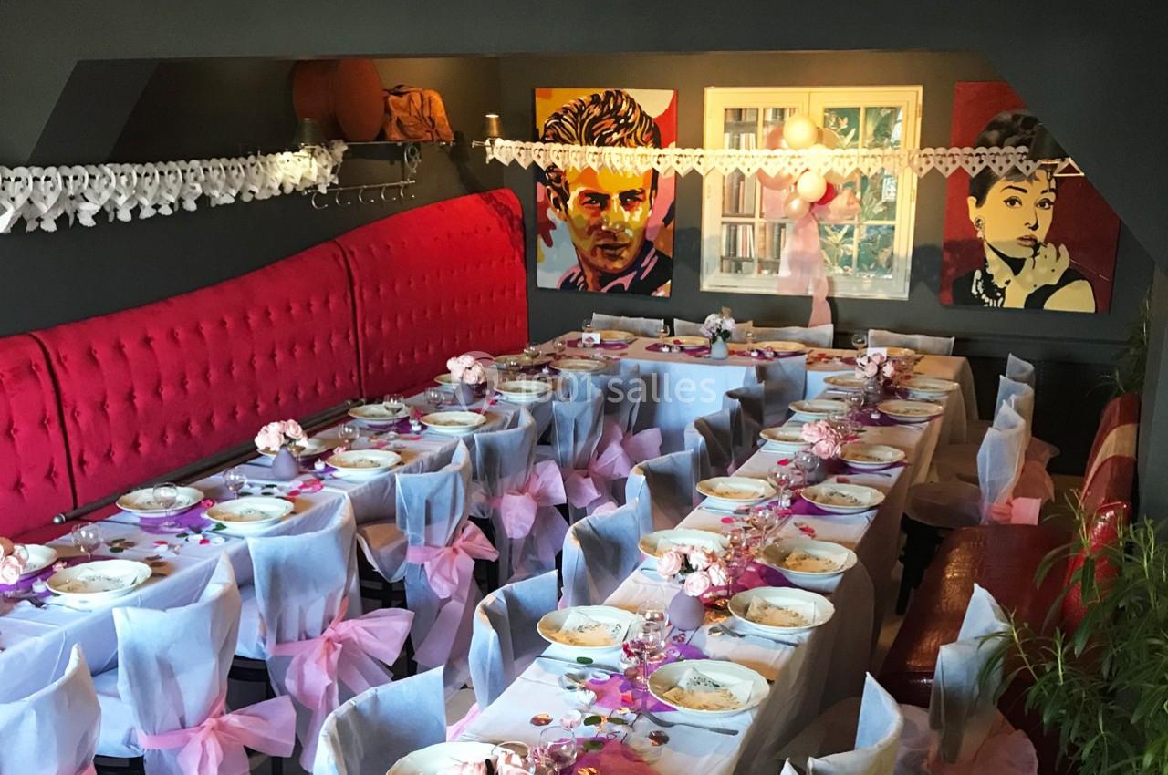 Salle décorée pour un événement avec longues tables dressées, nappes blanches, nœuds roses et portraits accrochés au mur.