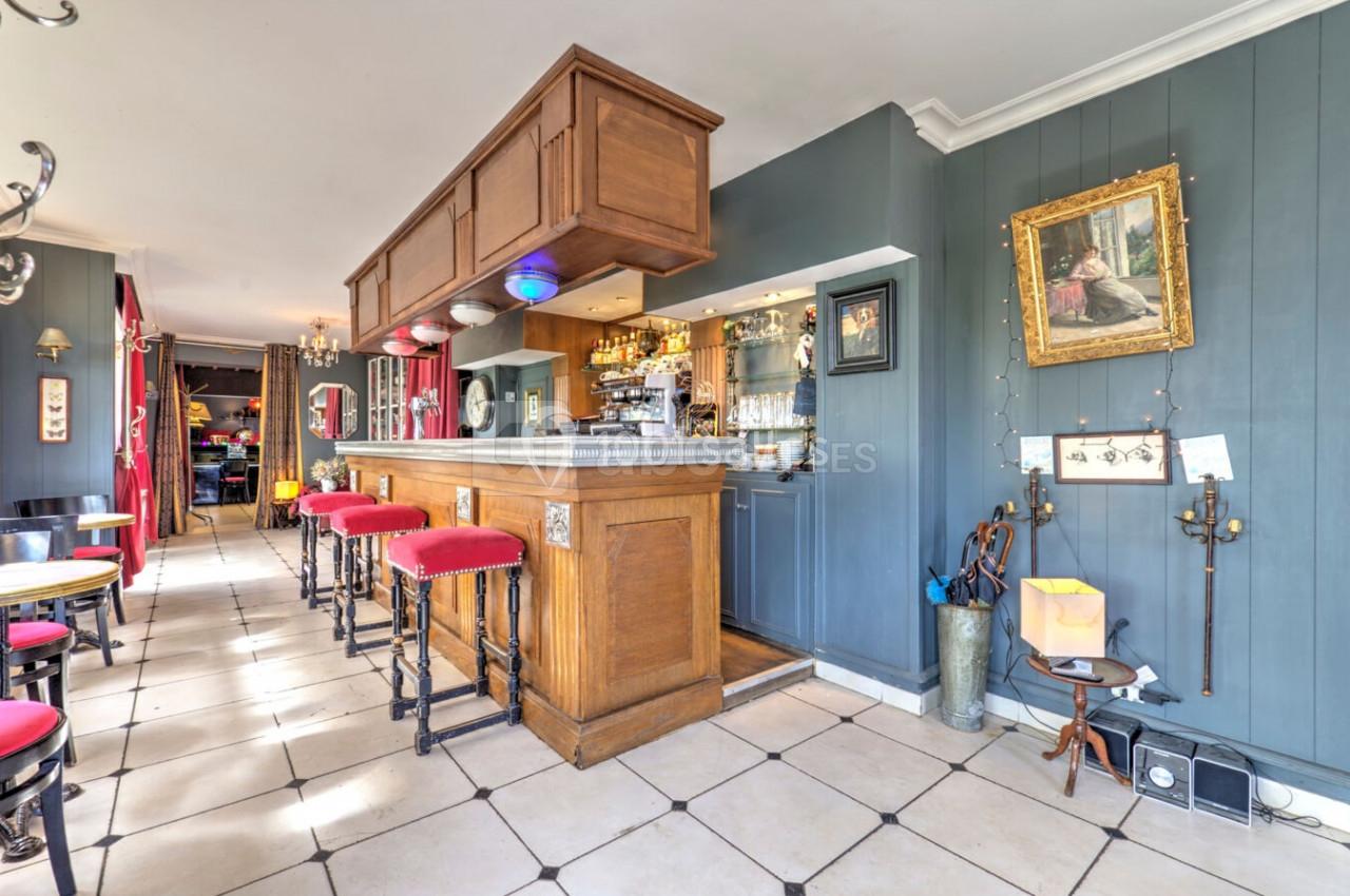 Intérieur d'un bar avec comptoir en bois, tabourets hauts, murs bleu-gris et décoration murale variée.