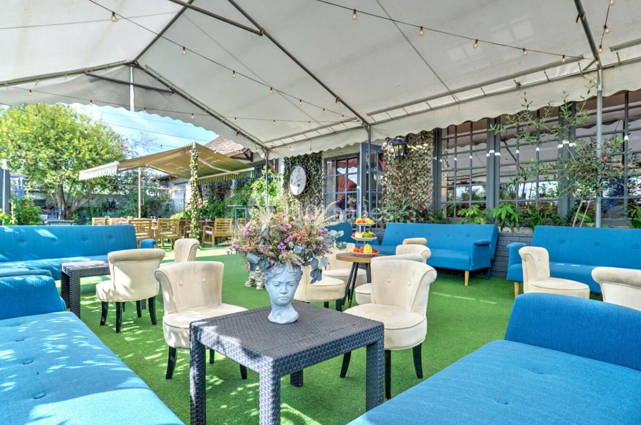 Terrasse aménagée avec des fauteuils bleus et beiges, tables en rotin, plantes et décoration florale sous une pergola.