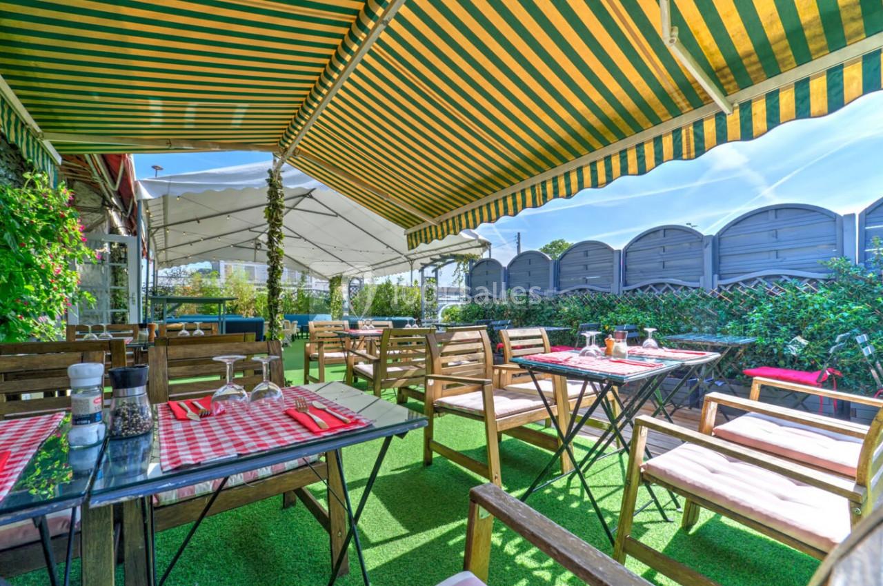 Terrasse extérieure avec tables en bois, nappes à carreaux rouges et blanches, sous des auvents rayés jaunes et verts.