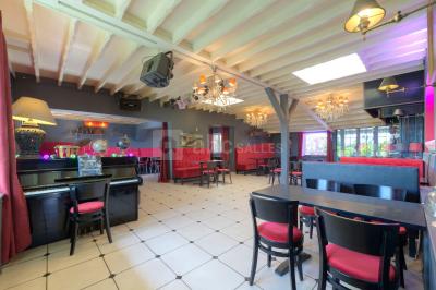 Salle décorée pour un événement avec longues tables dressées, nappes blanches, nœuds roses et portraits accrochés au mur.