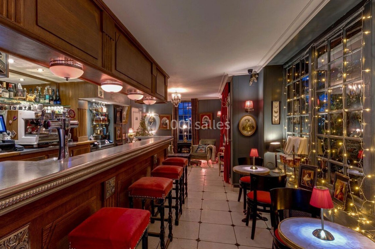 Intérieur d'un bar chaleureux avec comptoir en bois, tabourets rouges, tables éclairées et guirlandes lumineuses.