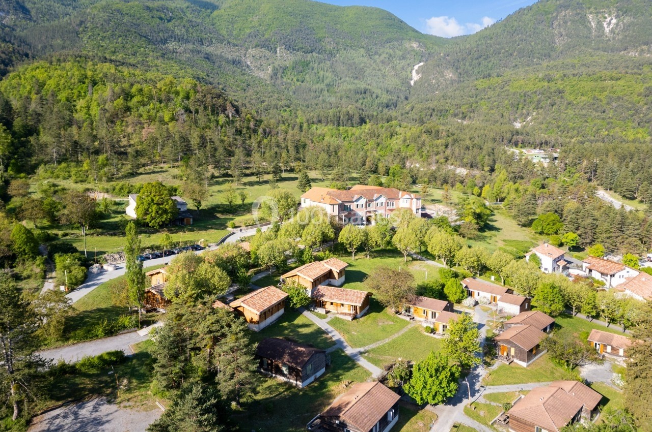 Vue aérienne d'un village de chalets entouré de verdure et de collines boisées sous un ciel bleu.