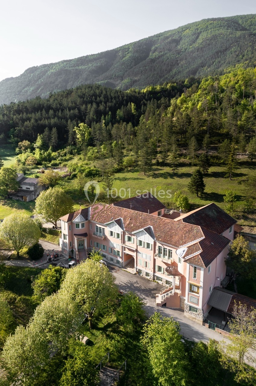 Vue aérienne d'un grand bâtiment rose entouré de verdure, avec une colline boisée en arrière-plan.