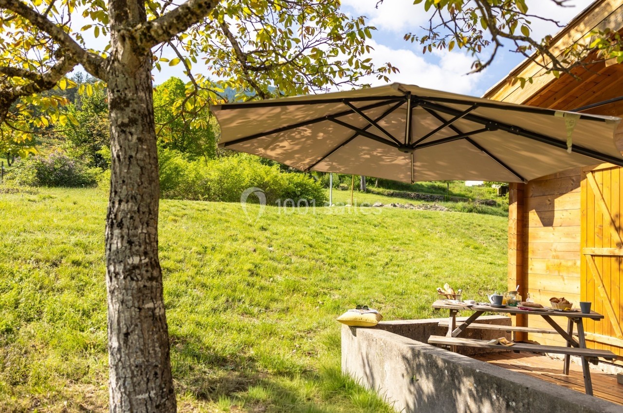 Table de pique-nique en bois sous un parasol beige, près d'un chalet en bois, entourée d'un paysage verdoyant.