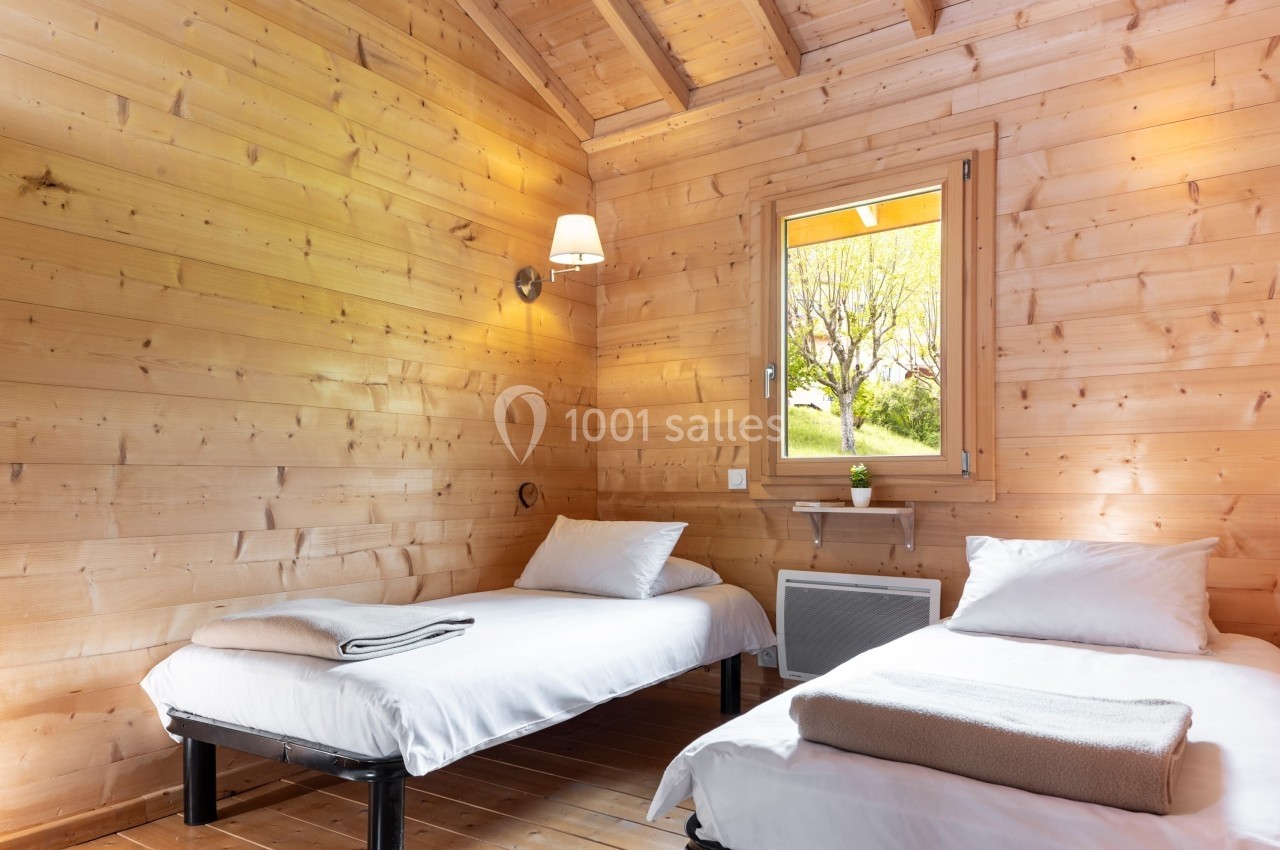 Chambre en bois avec deux lits simples, une fenêtre donnant sur un jardin et une applique murale allumée.