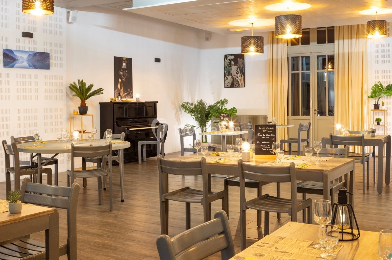 Salle de restaurant lumineuse avec tables en bois, chaises grises, piano noir et décorations végétales.