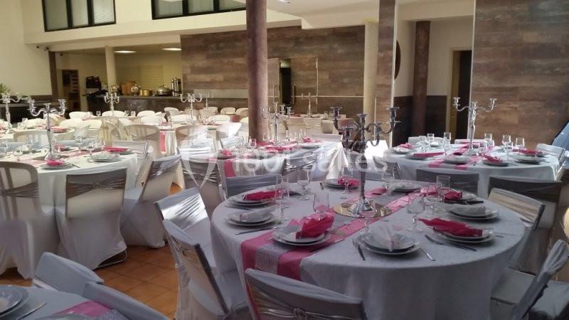 Salle de réception avec tables rondes dressées, nappes blanches, serviettes roses et chandeliers argentés.