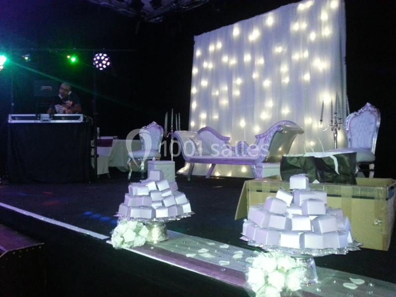 Scène décorée avec un canapé blanc, rideau lumineux, DJ en arrière-plan et pyramides de boîtes cadeaux au premier plan.