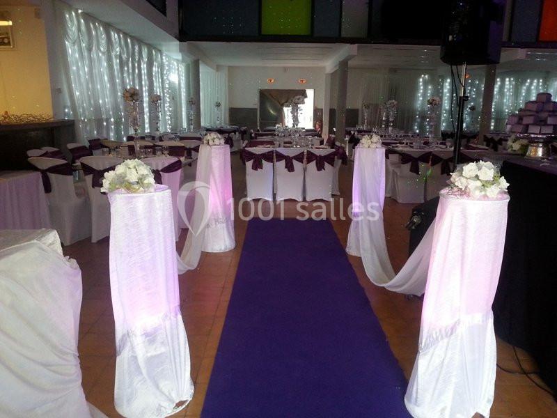 Salle de réception décorée avec des tables dressées, des housses blanches et des colonnes ornées de fleurs blanches.